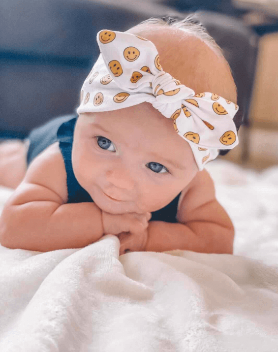 Baby girl is wearing a comfortable baby head wrap featuring a smiley pattern from By Bella Boutique. Baby girl is wearing a comfortable baby head wrap featuring a smiley pattern from By Bella Boutique.