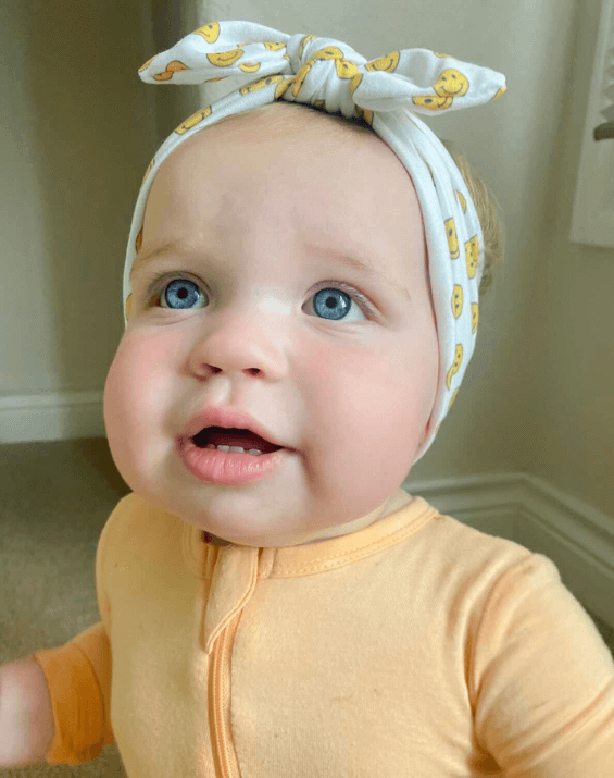 Baby girl is wearing a knotted head wrap featuring a smiley print from By Bella Boutique. Baby girl is wearing a knotted head wrap featuring a smiley print from By Bella Boutique.