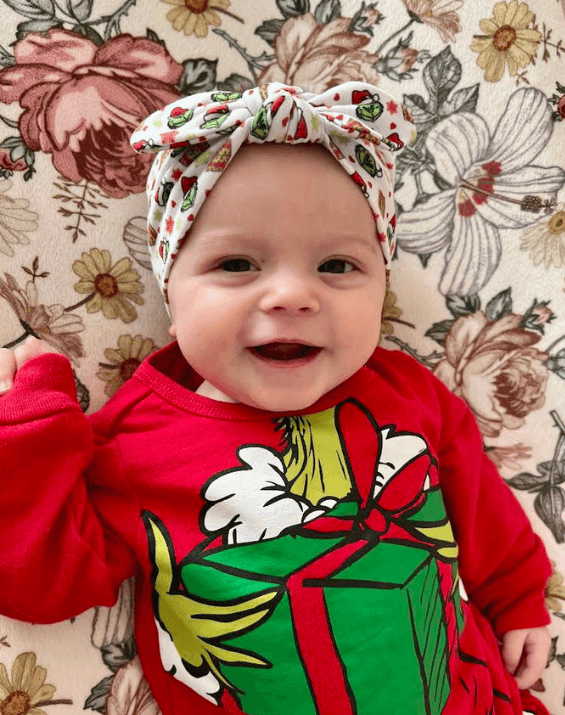 Baby girl is wearing a knot headband featuring a grinch pattern from By Bella Boutique.