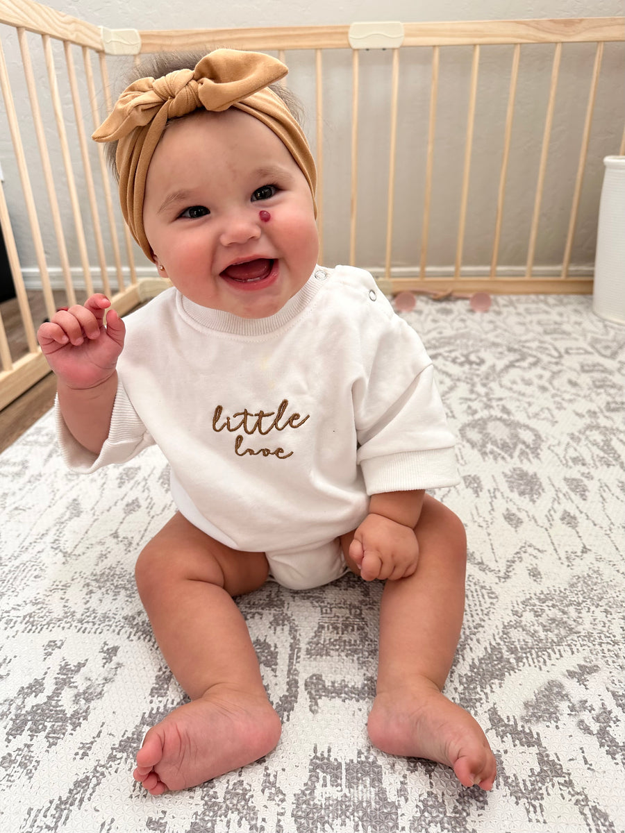 Baby wearing a mustard headband, smiling, sitting on a rug