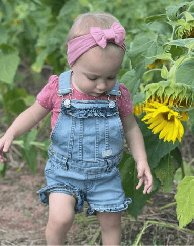 Little girl is standing in a sunflower field wearing a handmade pink bow headband from By Bella Boutique.